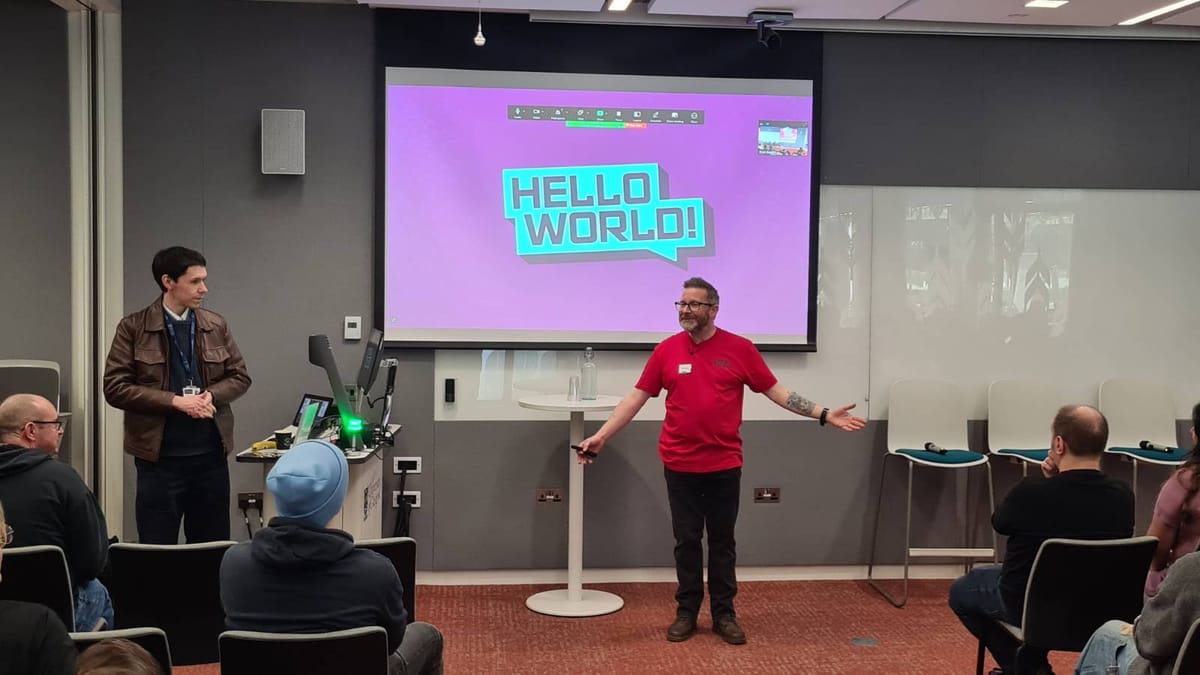 Man in red tshirt centre in frint of audience and urpleish screen saying hello Word to left is another gent standing looking on